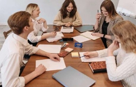 young-professional-service-team-meeting-collaboration A group of five young professional colleagues sitting around a large wooden conference table, smiling and collaborating during a team meeting.